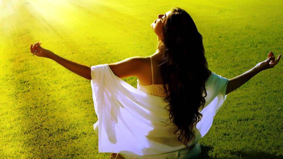 Frau genießt Sonnenlicht auf der Wiese, Arme ausgebreitet, entspannte Pose.