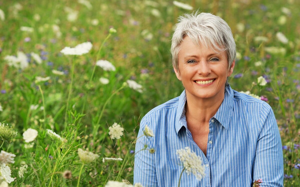 Ältere Frau mit grauem Haar lächelt in einer blühenden Wiese.