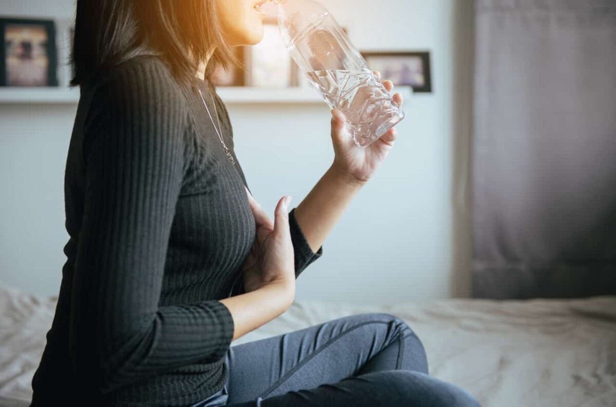 Frau in schwarzem Oberteil trinkt aus einer klaren Wasserflasche im Schlafzimmer.