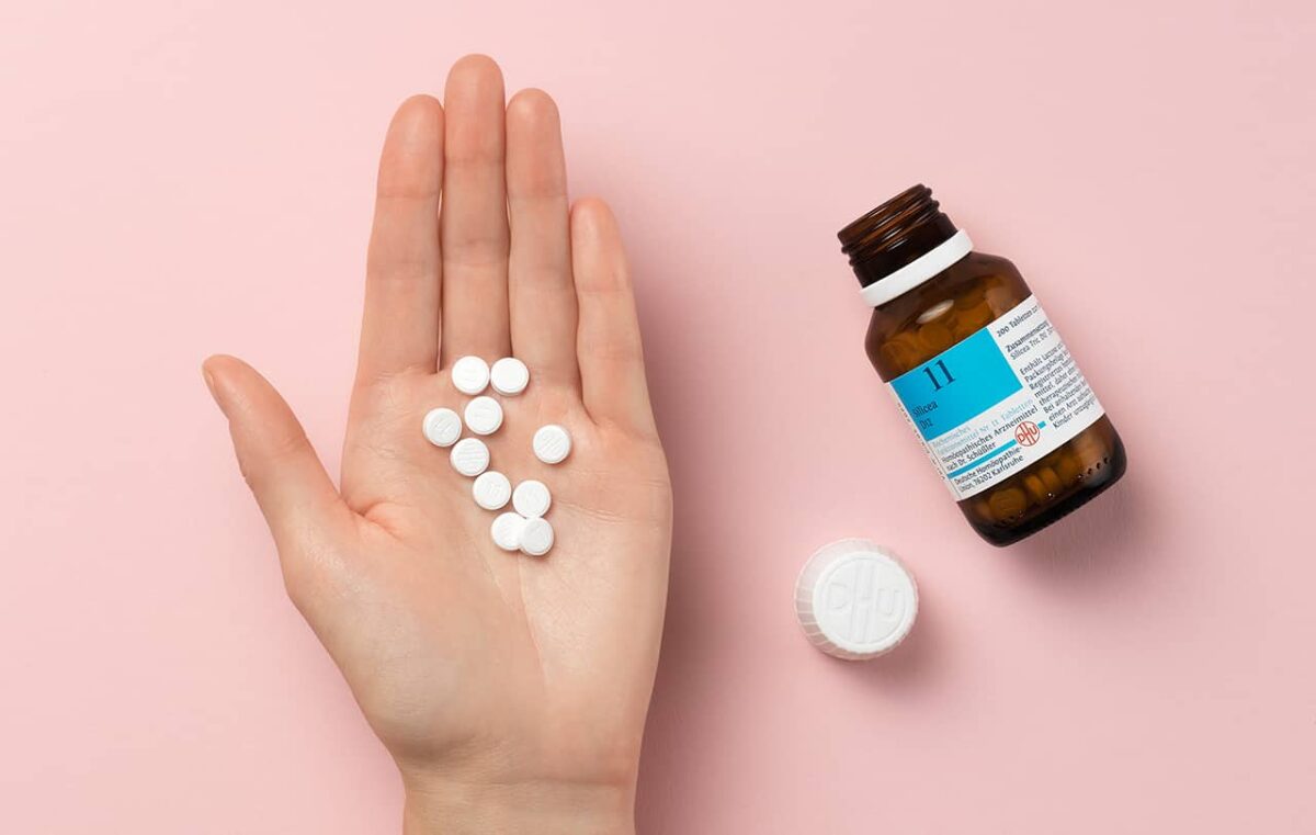 Hand mit weißen Tabletten und brauner Pillendose auf rosa Hintergrund.