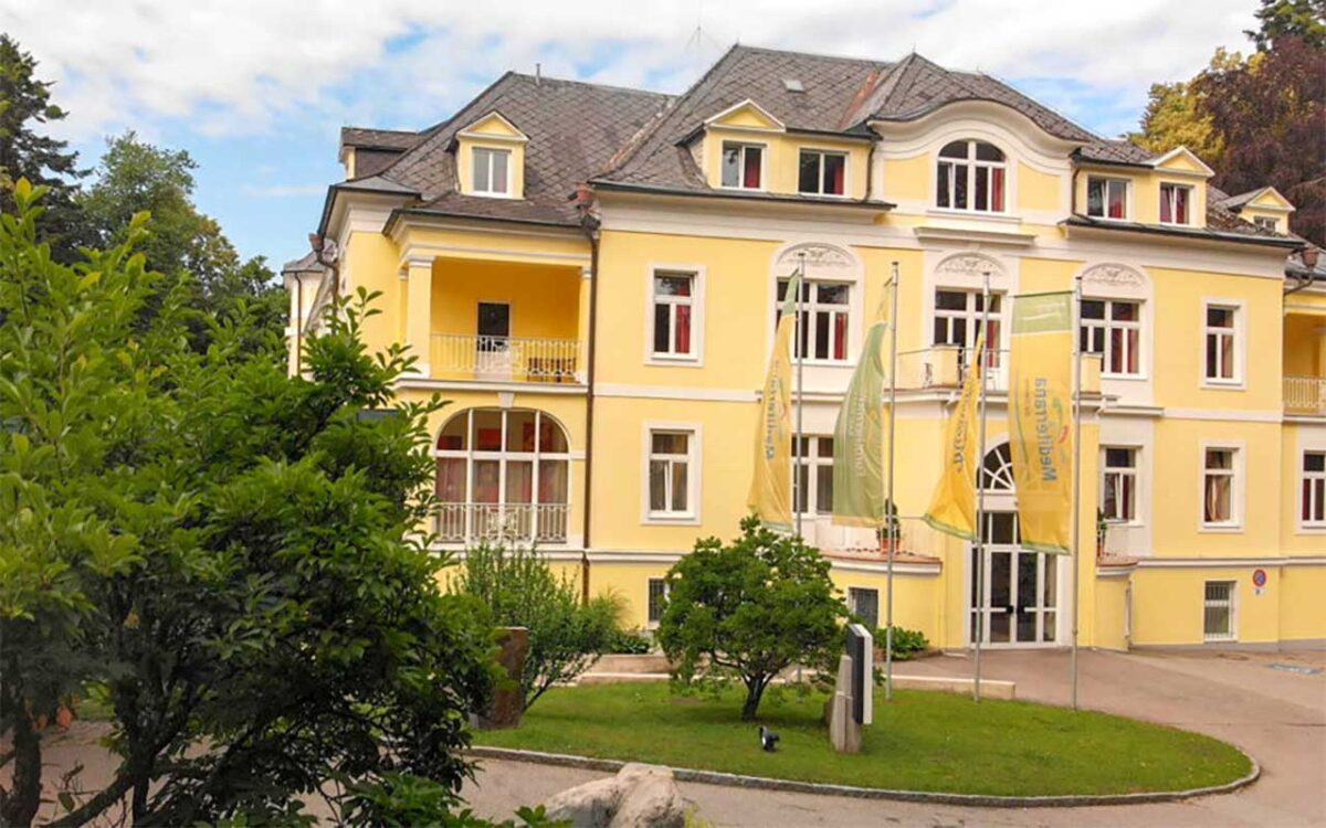Gelbe historische Villa mit Flaggen, umgeben von grüner Landschaft und klarem Himmel.